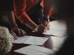 Como imagen destacada para este texto sobre el pago de Lo que debes saber de las regalías de franquicias hemos seleccionado una fotografía en la que se aprecian la senior un par de brazos que debajo tienen una mesa de manera, sobre ella posan una hojas blancas. La mano derecha firma una de la hoja, mientras otra mano se recarga. Junto a ellas parecen otras dos que se posan sobre otra hoja.