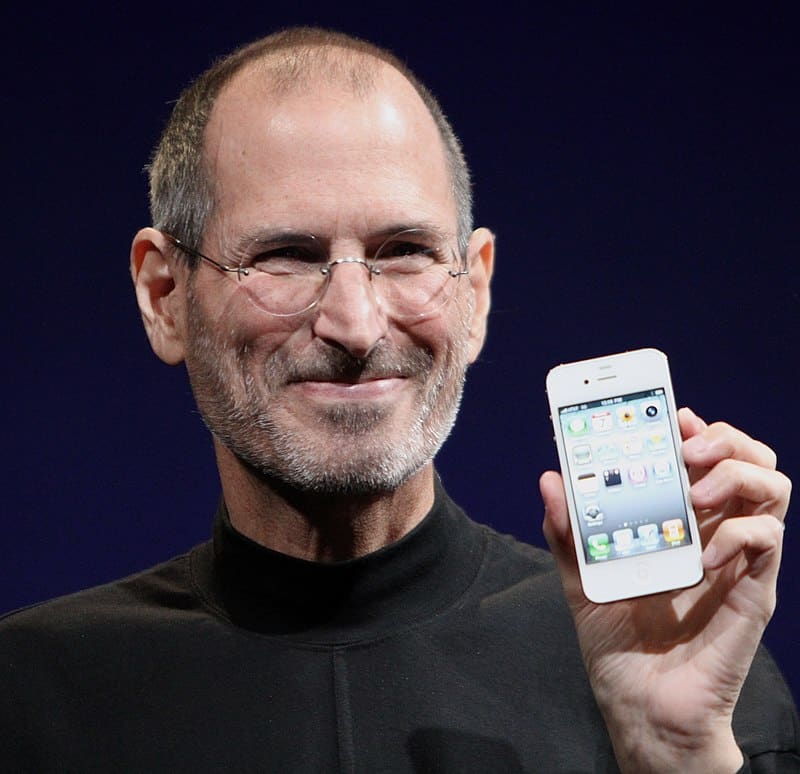 El emprendedor Steve Jobs aparece en esta fotografía a color, viendo de frente. Es un hombre sonriente con barba corta y blanca. Viste cuello alto color negro y lentes oftalmológicos discretos. Con la mano izquierda sostiene un iPhone prendido en la apantalla de inicio hacia la cámara.