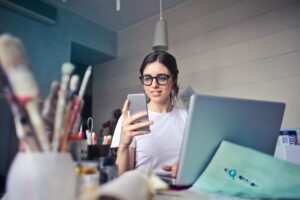 Como imagen destacada para este texto sobre como alcanzar tu independencia financiera hemos seleccionado la fotografía de una mujer joven, queden esta sentada y tiene un celular en la mano una computadora portátil al frente. Además, sobre la mesa se perciben pinceles colocados en contenedores.