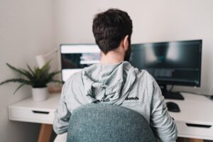 Como imagen destacada de este texto de consejos para triunfar en el trabajo remoto o home office, tenemos una fotografía en la que vemos a un hombre de espaldas, quien esta sentado frente a dos monitores para trabajar en casa. Los monitores posan sobre un escritorio blanco, amplio en el que también se ve una pequeña maceta blanca en la que hay un helecho.