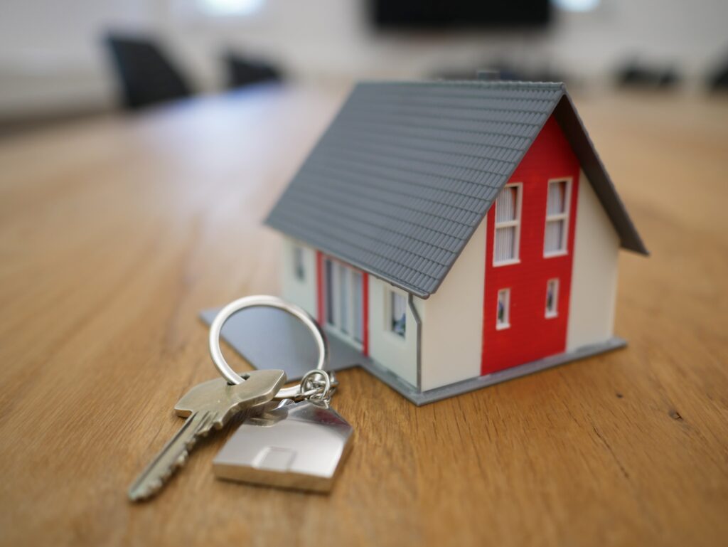 La imagen destacada de este texto sobre qué es una hipoteca y cómo funciona es una fotografía de la maqueta de una casa que solo se ve su exterior, sobre una mesa de madera y junto a unas una llave con un llavero. la casa tiene un tejado gris y sus paredes son rojas y crema.
