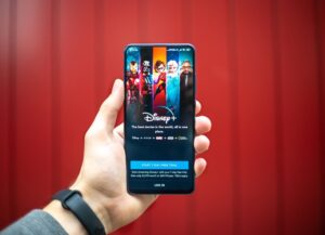 Como imagen destacada de esta nota sobre el despidos de Peter Rice, ejecutivo de Disney, tenemos una fotografía de un la aplican para streaming de la firma, en un celular que es sostenido por una mano. Al Fondo se simula un telón rojo.