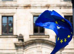 Como imagen destacada para este texto sobre el Sistema Europeo de Información y Autorización de Viajes (ETIAS) tenemos una imagen destacada en la que se ve la bandera de la Unión Europea (EU) ondeando en el lado derecho. Como fondo del símbolo de esta unión de Europa, tenemos un edificio.