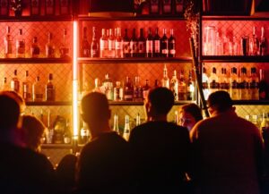 En este texto sobre los derechos de los consumidores restaurantes, bares y cantinas, tenemos como imagen destacada una fotografía de la barra de un bar, donde vemos a varias personas de espalda y las repisas donde están las botellas iluminadas con luces de colores rosas, amarillas y naranjas.