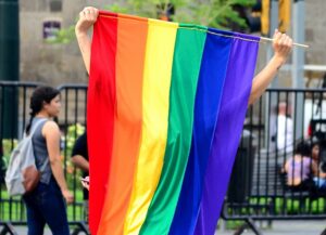 Como imagen destacada sobre esta nota de los avances en materia de diversidad sexual para proteger a la comunidad LGBTQ+ en México, tenemos una fotografía en la que resalta al centro una bandera gay sostenida por una persona quien no se ve pues la coloca extended en su espalda.