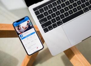 Como imagen destacada para esta nota titulada: Ganancias de Meta caen 36%, tenemos una fotografía en la que vemos desde arriba a un celular sobre una mesa de cristal, junto a una computadora portátil. El celular muestra en su pantalla estar abierta la aplicación de Facebook, marca insignia de la compañía.