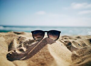 Para este texto sobre tips para lograr un verano sin derroche, frente a los gastos de la temporada, tenemos una imagen destacada que es una fotografía de una gafas de sol sobre la arena, frente al mar de día.